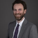 Professional man in a gray suit with a purple tie smiling at the camera.