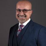 Smiling man in a suit and tie with glasses against a dark background.