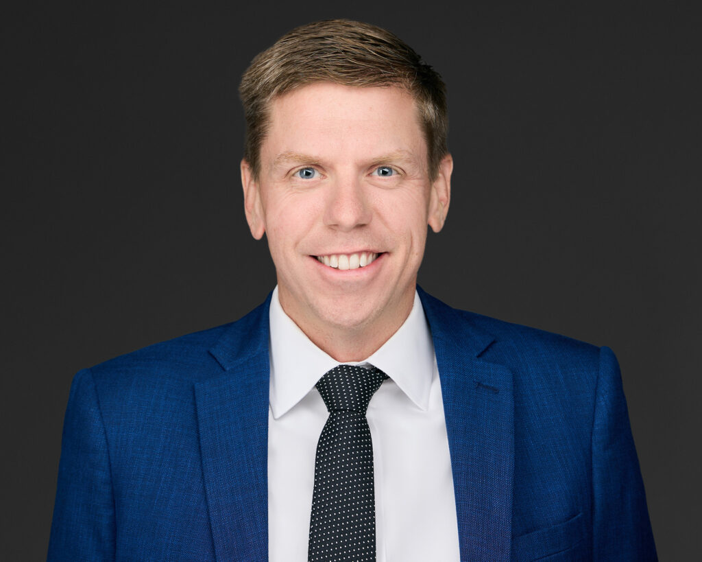 Professional man in blue suit and tie smiling against dark background.