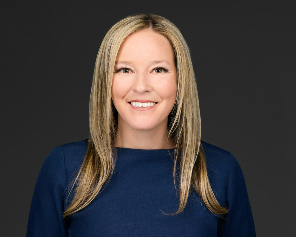 Smiling woman with long blonde hair wearing a blue top against a dark background.