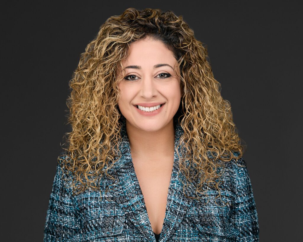 Smiling woman with curly hair in a blue patterned jacket against a dark background.