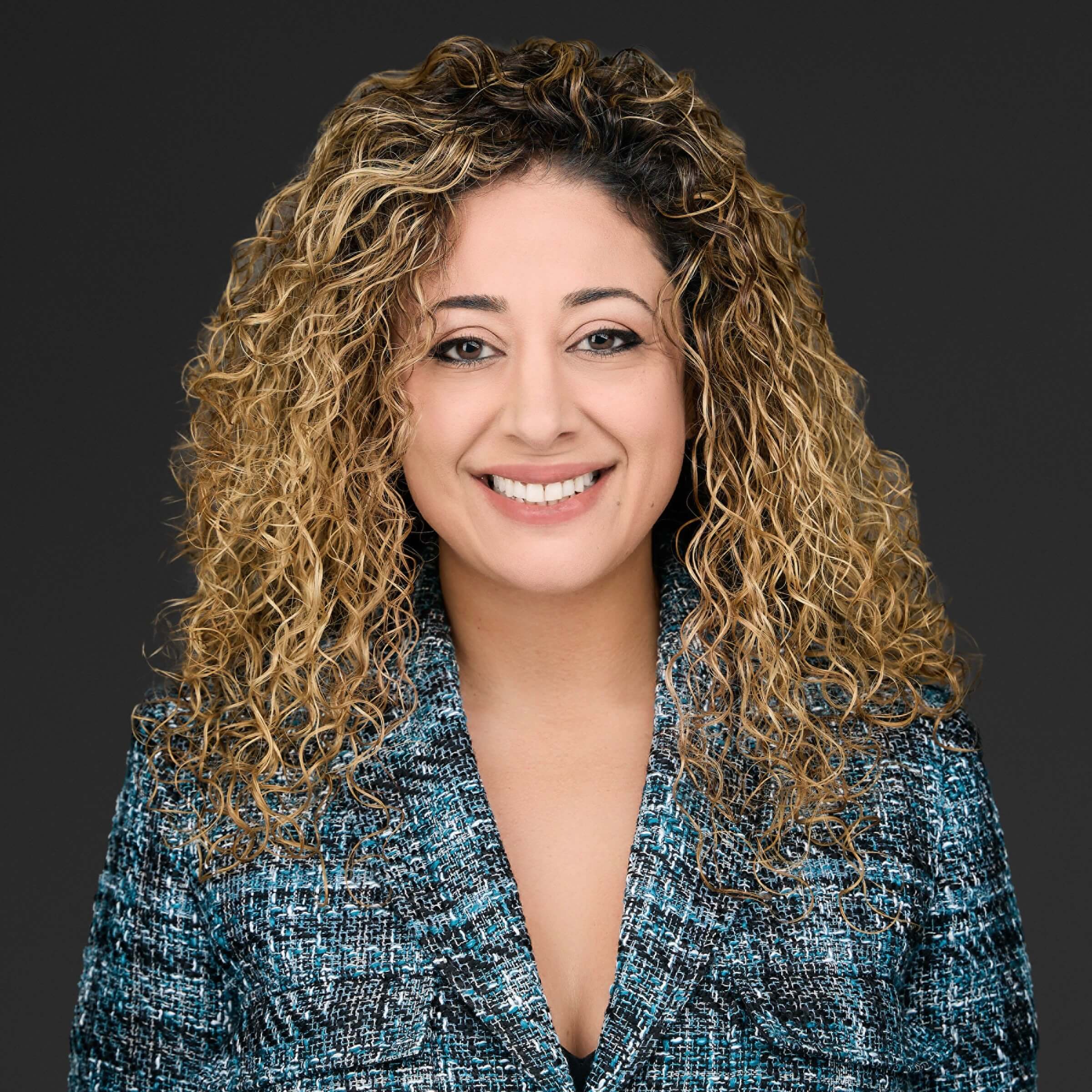 Smiling woman with curly hair in a blue patterned blazer against a dark background.