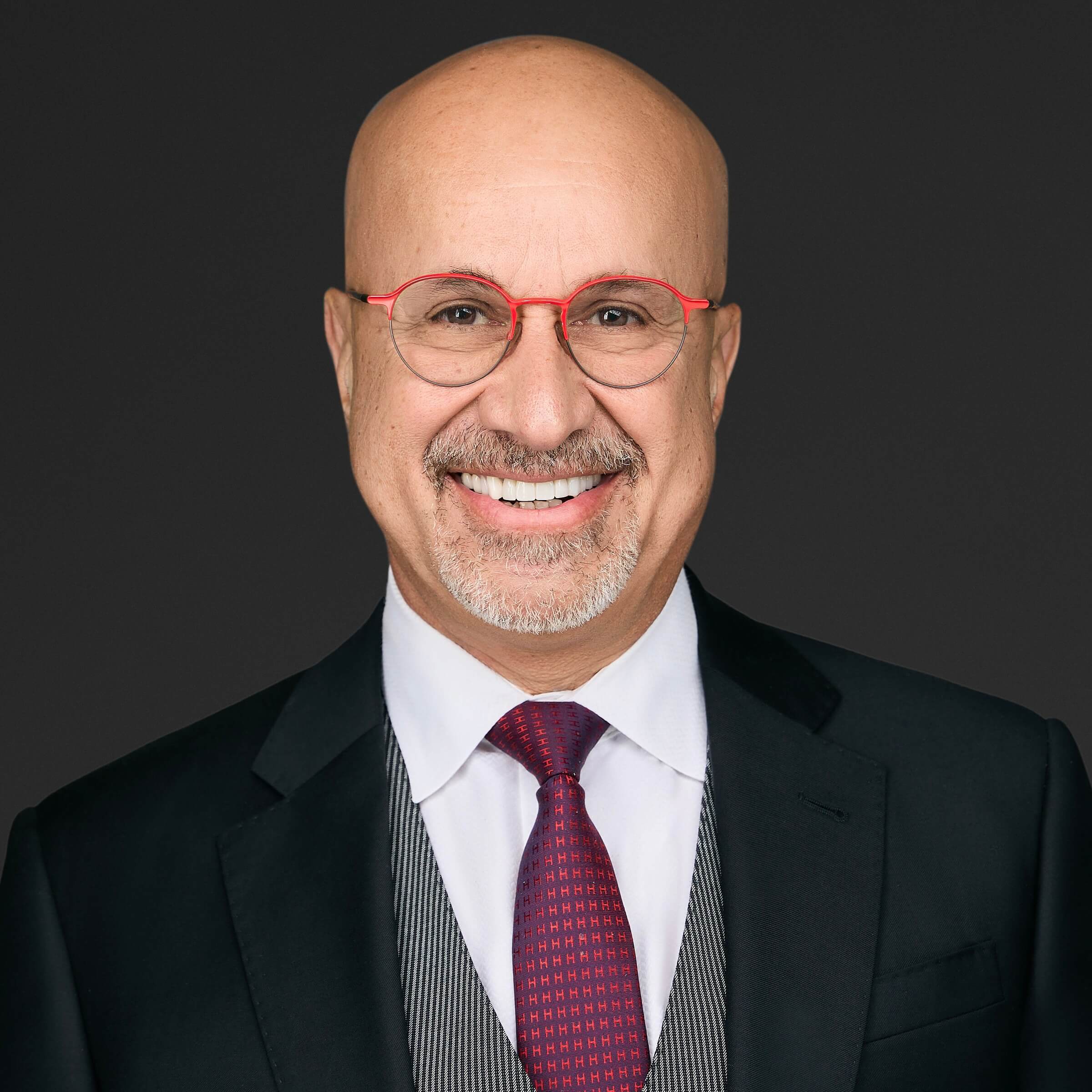 Smiling man in a suit and red glasses against a dark background.