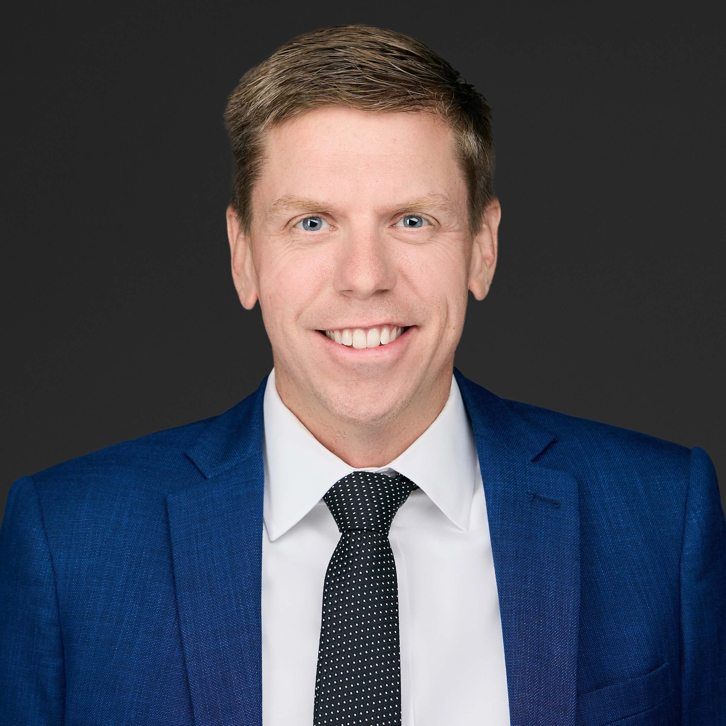 Professional man in blue suit smiling against dark background.