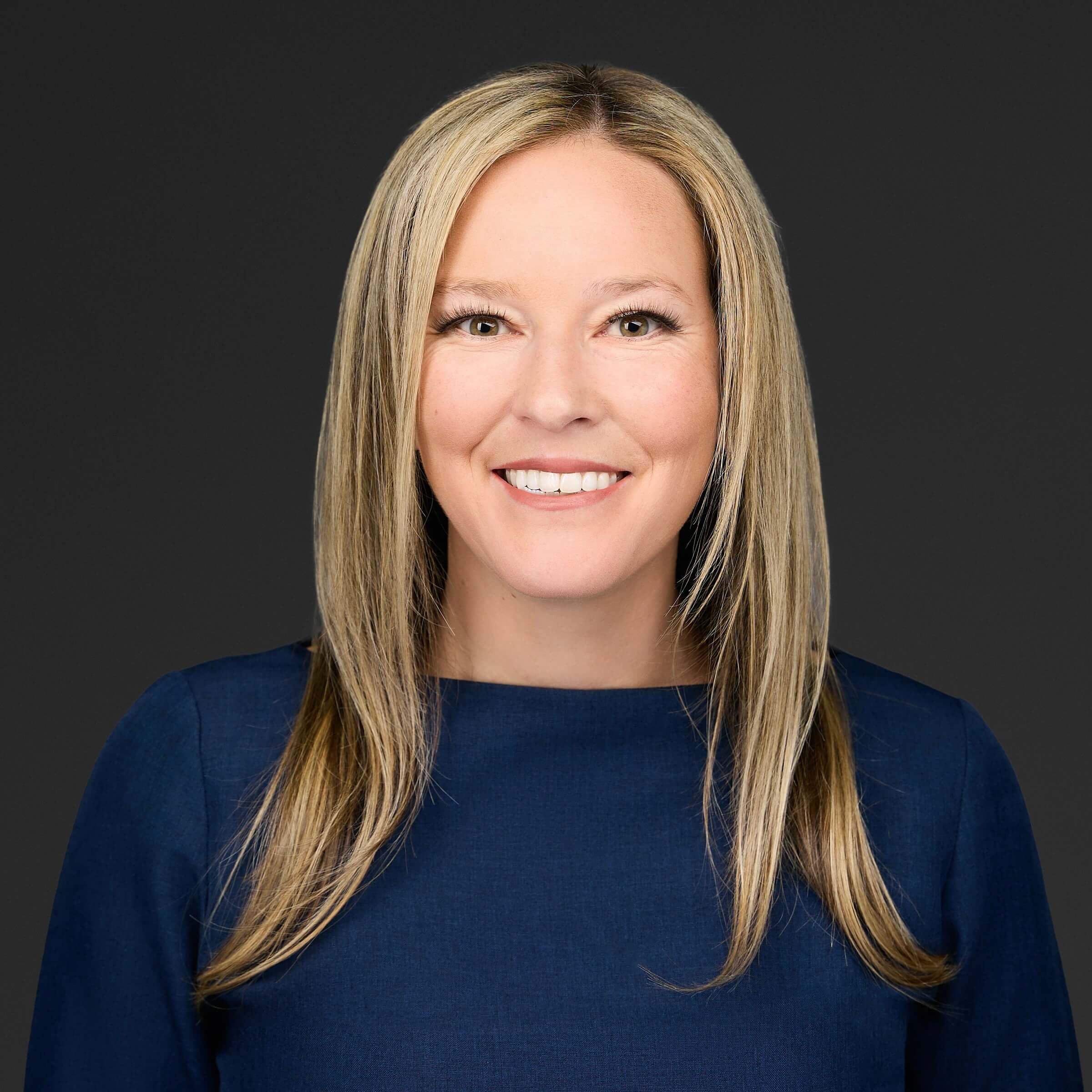 Smiling woman with long blonde hair wearing a navy blue top against a dark background.